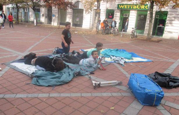 Eine Gruppe von Kindern spielt auf einer Decke auf einem Platz vor einem Wettbüro.