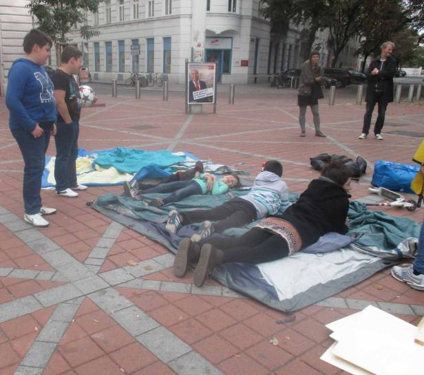 Eine Gruppe von Menschen liegt auf Matten auf einem Platz.