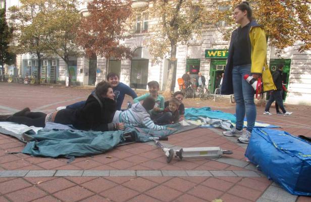 Eine Gruppe von Kindern und eine Frau liegen auf einer Plane auf einem Platz.