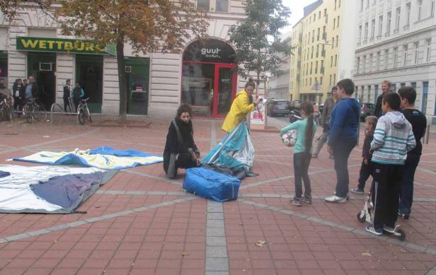 Zwei Frauen bauen ein Zelt auf einem Platz auf, während eine Gruppe Kinder zusieht.