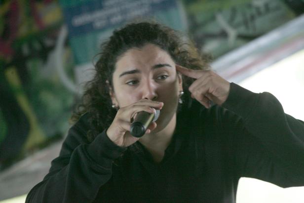 Eine Frau mit lockigem Haar rappt in ein Mikrofon und tippt sich an die Stirn.