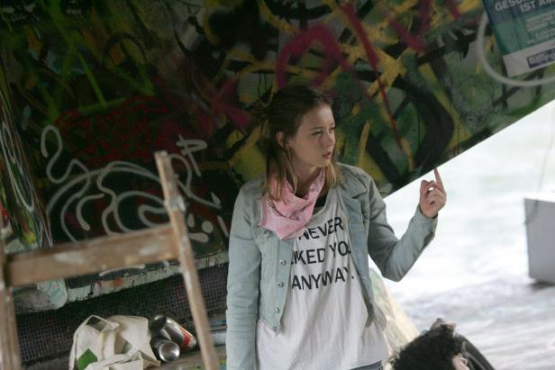 Eine junge Frau steht vor einer Wand mit Graffiti und gestikuliert.