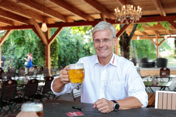 Ein Mann mit Brille prostet mit einem Bierkrug in einem Biergarten.