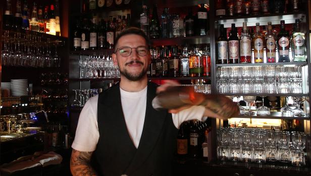 Ein Barkeeper mit Tätowierungen mixt einen Cocktail vor einem Regal voller Spirituosen und Gläser.