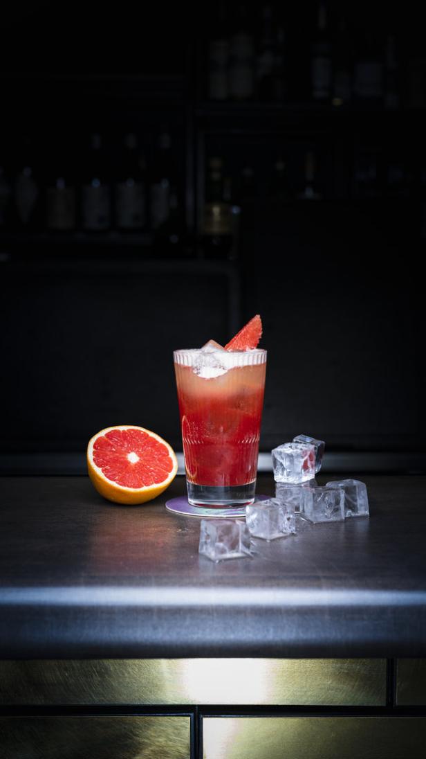 Ein Glas mit einem roten Cocktail, Eiswürfeln und einer halben Grapefruit auf einer Bar.