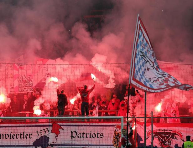 Fußballfans zünden rote Bengalos hinter einem Zaun mit einem Vereinsbanner.