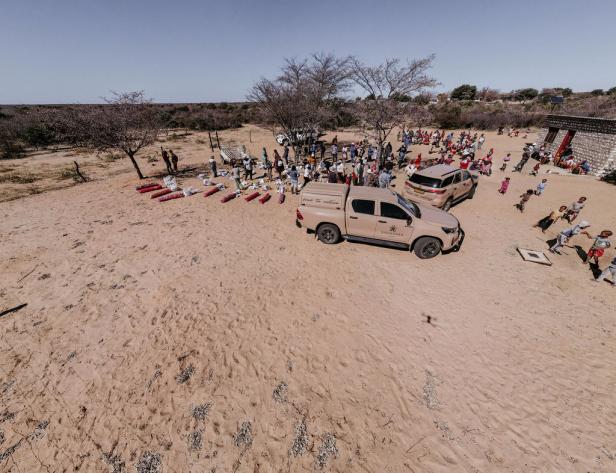 Eine Gruppe von Menschen und Kindern versammelt sich in einer sandigen Landschaft mit zwei hellbraunen Geländewagen.