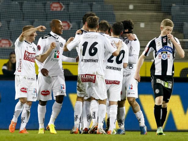 Eine Gruppe Fußballspieler in weißen Trikots umarmt sich, während ein Spieler in Schwarz-Weiß traurig daneben steht.