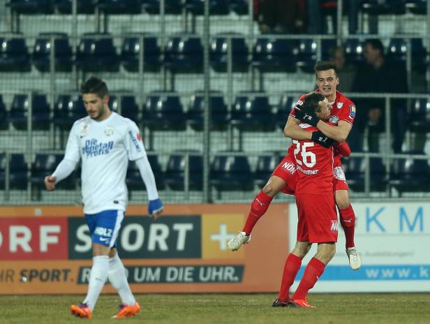 Zwei Fußballspieler in roten Trikots umarmen sich auf dem Spielfeld, während ein anderer Spieler in Weiß vorbeigeht.