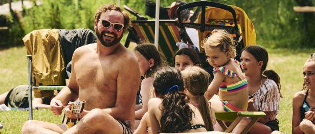 Ein Mann mit Ukulele sitzt barfuß mit einer Gruppe von Kindern im Freien.