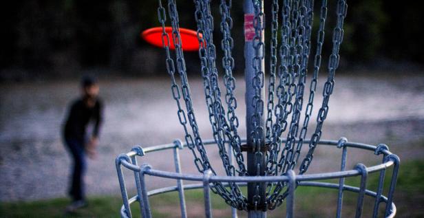 Ein Discgolf-Korb mit Ketten und ein Spieler im Hintergrund wirft eine Scheibe.