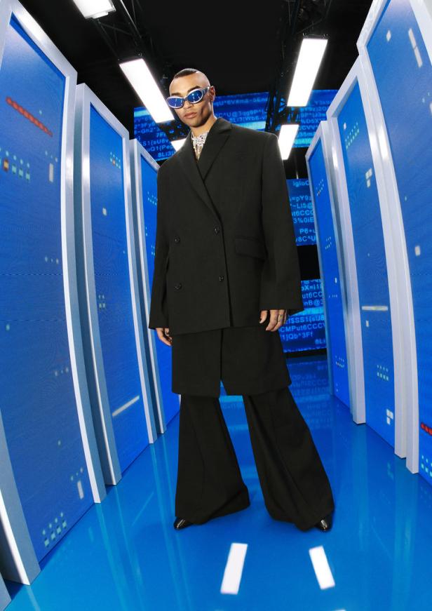 Ein Mann mit Sonnenbrille trägt einen übergroßen schwarzen Anzug vor einem blauen Hintergrund mit Computercode.