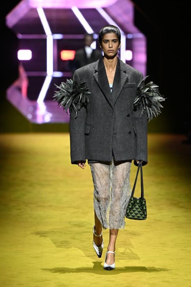Ein Model auf dem Laufsteg, gekleidet in einen übergroßen Blazer mit Federapplikationen und einem schimmernden Rock.