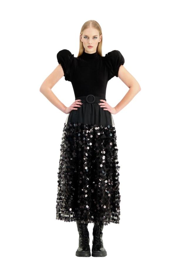 Eine Frau in einem schwarzen Kleid mit Paillettenblumen und Stiefeln posiert vor einem weißen Hintergrund.