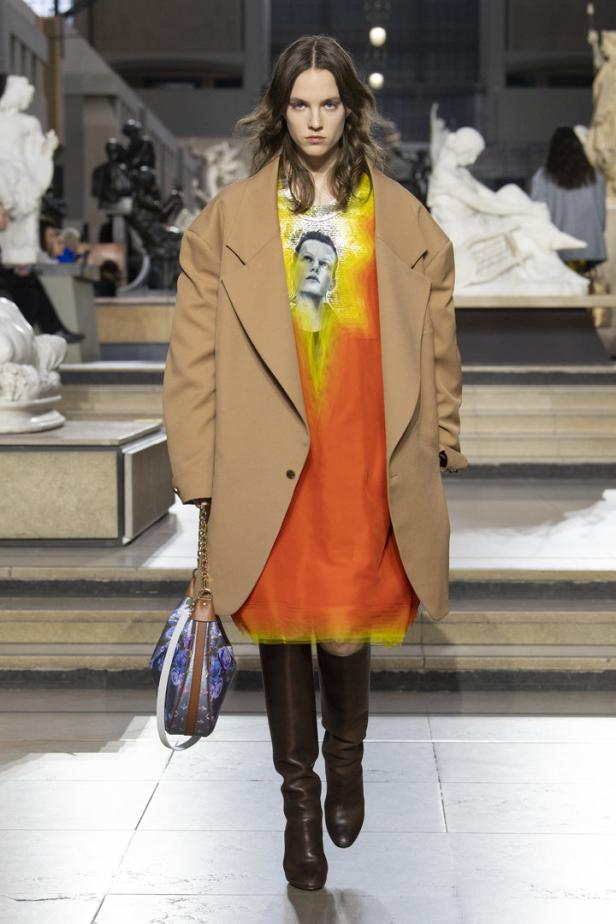 Eine Frau in einem orangefarbenen Kleid, einem beigen Blazer und braunen Stiefeln auf einem Laufsteg.