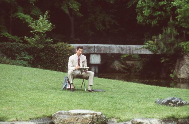 Prinz Charles sitzt auf einer Wiese und malt mit Blick auf eine Steinbrücke.