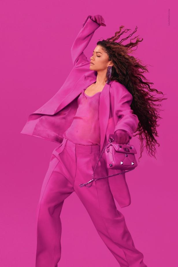 Zendaya trägt einen pinkfarbenen Hosenanzug und eine passende Handtasche vor einem pinkfarbenen Hintergrund.
