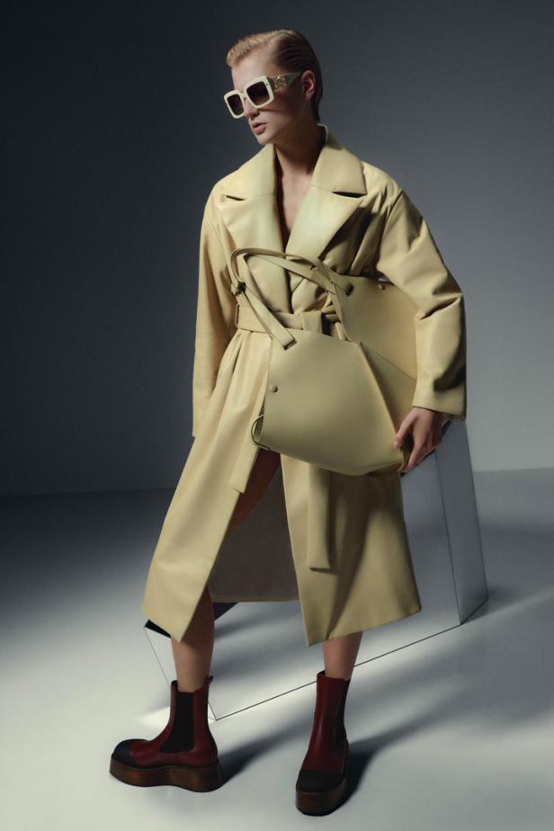 Eine Frau mit Sonnenbrille, Trenchcoat und Handtasche posiert vor einem grauen Hintergrund.