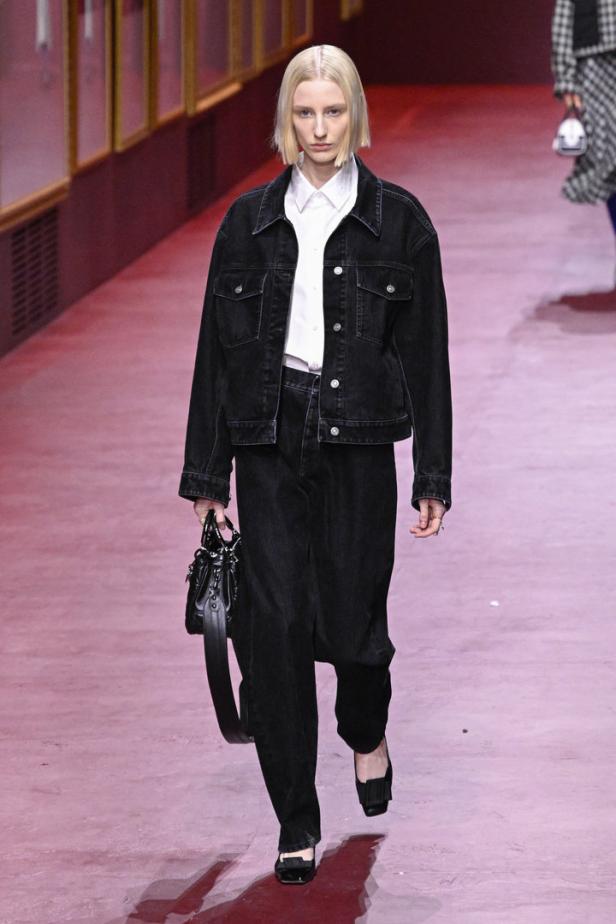 Eine Frau mit blondem Bob trägt ein schwarzes Jeans-Outfit und eine Handtasche auf dem Laufsteg.