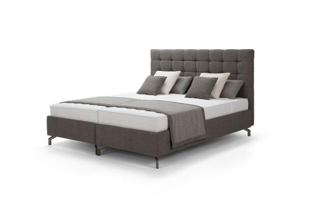 Ein Boxspringbett mit grauem Stoffbezug, Kopfkissen und einer Tagesdecke.