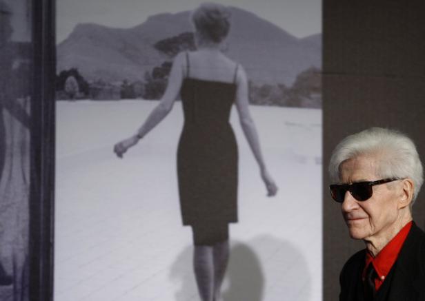 Ein älterer Mann mit Sonnenbrille steht vor einem Schwarzweißbild einer Frau in einem Kleid.