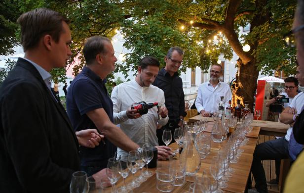 Eine Gruppe von Menschen steht um einen Tisch mit Weingläsern und einer Flasche Wein.