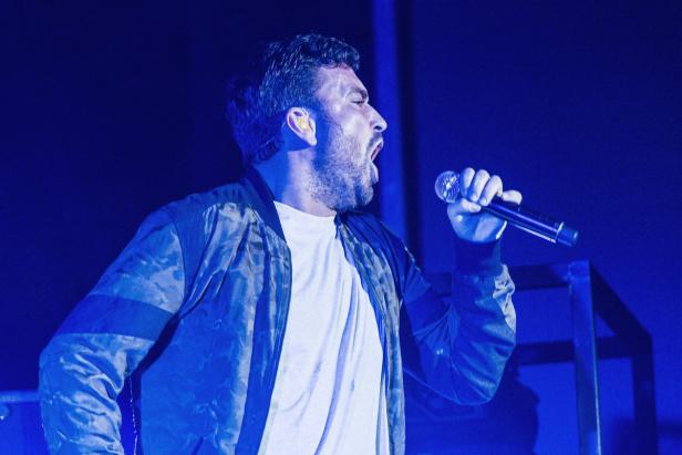 Ein Mann mit Bart singt in ein Mikrofon auf einer Bühne mit blauem Licht.