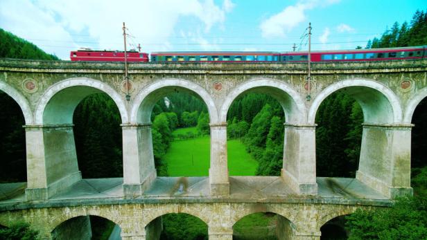 Ein roter Zug fährt über eine alte Steinbogenbrücke.