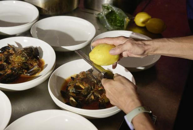 Eine Person reibt Zitronenschale über eine Schüssel mit Muscheln in Tomatensauce.
