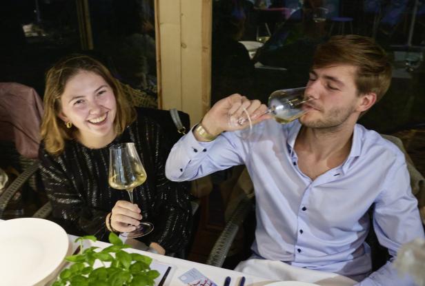 Eine junge Frau lächelt, während ein junger Mann Weißwein trinkt.