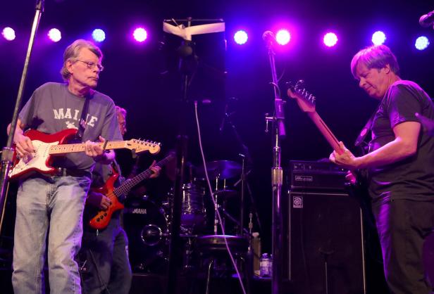 Stephen King spielt Gitarre auf einer Bühne mit lila Beleuchtung.