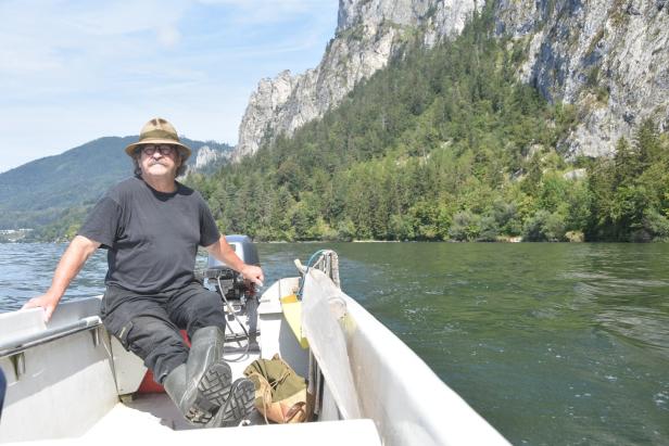 Ein Mann mit Hut fährt mit einem kleinen Motorboot auf einem Fluss entlang einer bewaldeten Felswand.