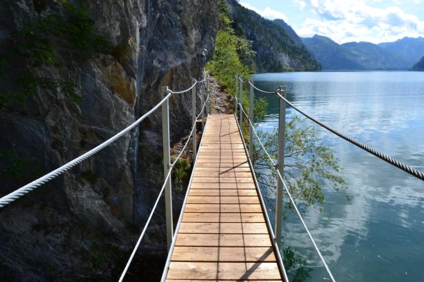 Ein schmaler Holzsteg führt an einer Felswand entlang über einen See.