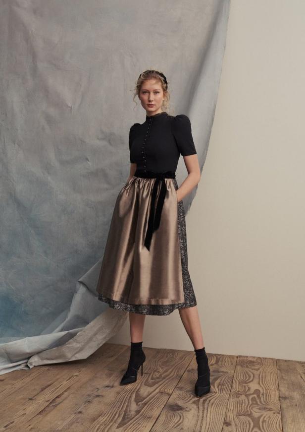 Eine Frau in einem Dirndl mit schwarzer Bluse und goldenem Rock mit schwarzer Schleife und schwarzen Stiefeln.