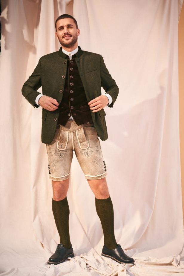 Ein Mann in Tracht mit Lederhose, Weste und Jacke vor einem hellen Hintergrund.