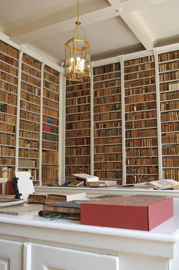 Ein Raum mit deckenhohen Bücherregalen und einem Schreibtisch mit Büchern und einer Lampe.