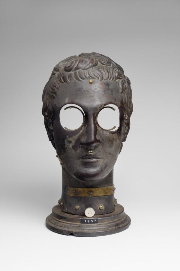 Eine metallene Gesichtsmaske mit ausgeschnittenen Augenlöchern auf einem Sockel.