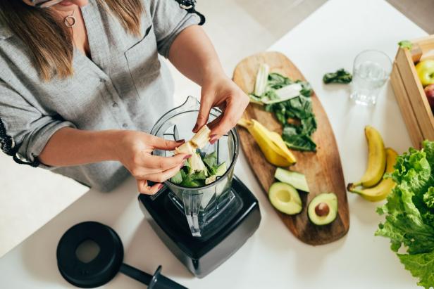 Eine Frau gibt Bananenstücke in einen Mixer mit grünem Gemüse.