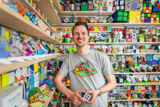 Ein Mann steht lächelnd vor Regalen voller Rubik's Cubes und hält einen in der Hand.