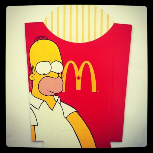 Homer Simpson vor einer McDonald's-Pommes-Verpackung mit kanadischem Logo.