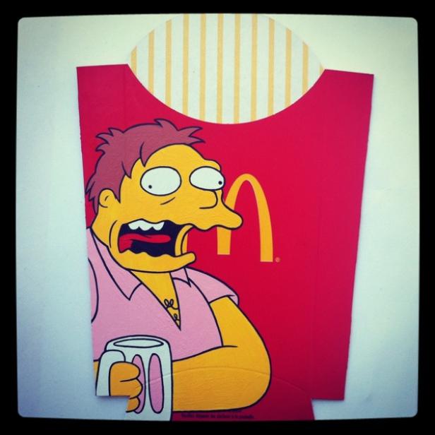 Ein Comic-Bild von Barney Gumble vor einer McDonald's-Pommes-Verpackung mit einem Bierkrug in der Hand.