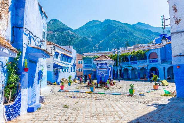 Ein belebter Platz in Chefchaouen, Marokko, mit blau gestrichenen Häusern und Bergen im Hintergrund.