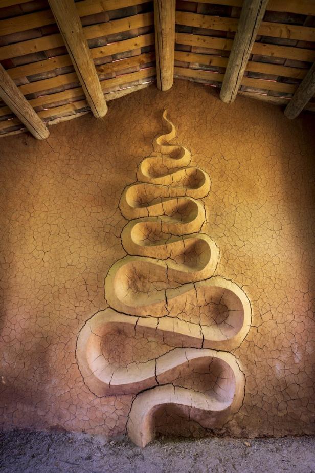 Eine spiralförmige Skulptur an einer Lehmmauer unter einem Holzdach.