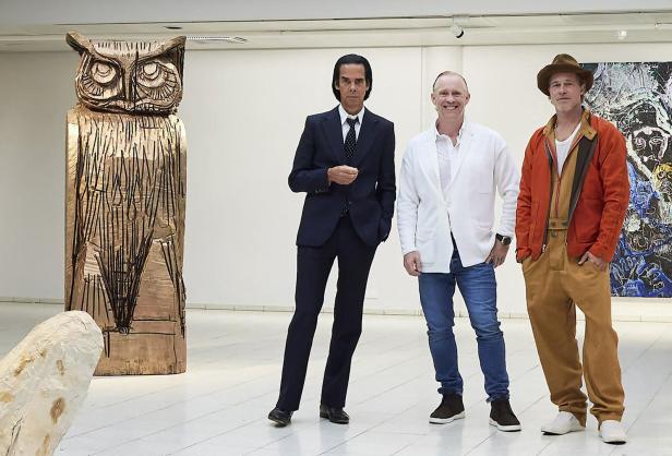 Nick Cave, Thomas Houseago und Brad Pitt posieren vor einer Eulen-Skulptur in einer Kunstgalerie.