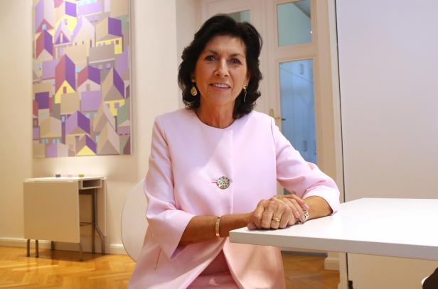 Eine Frau in einem rosa Blazer sitzt an einem Tisch vor einem abstrakten Gemälde.