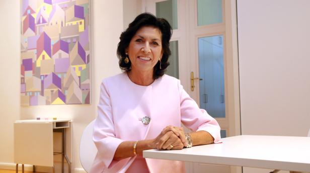 Eine lächelnde Frau in einem rosa Blazer sitzt an einem Tisch.