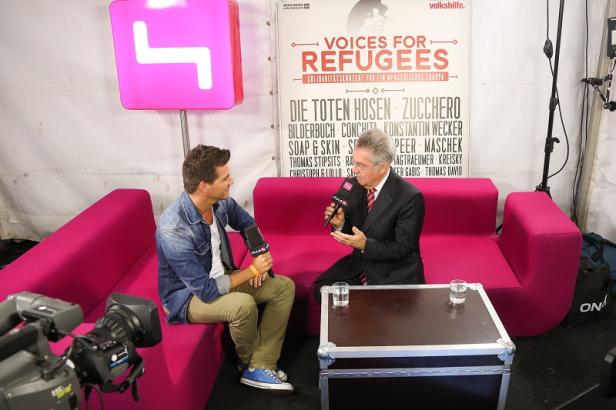 Zwei Männer sitzen auf einem pinkfarbenen Sofa und führen ein Interview vor einem „Voices for Refugees“-Plakat.