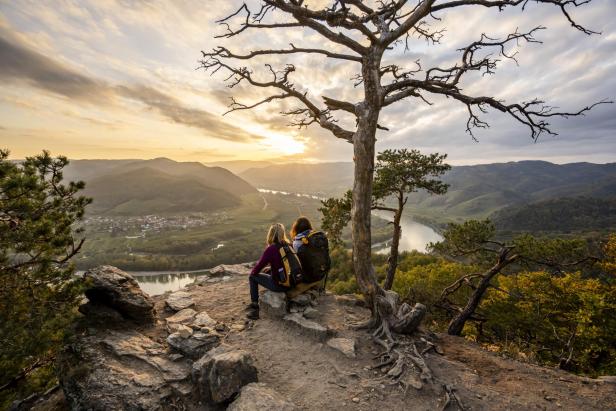 Zwei Wanderer sitzen auf einem Felsen und blicken auf eine Flussschleife im Tal bei Sonnenuntergang.
