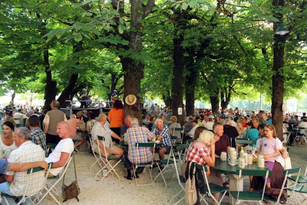 Viele Menschen sitzen an Tischen unter Bäumen in einem Biergarten.
