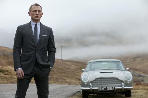 Daniel Craig als James Bond posiert vor einem Aston Martin in einer nebligen Landschaft.
